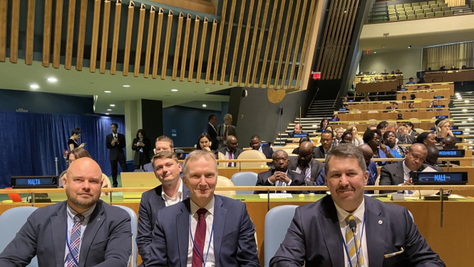 Trīs vīrieši, ģērbušies uzvalkos sēž pie koka galda ar zaļu pārklāju un skatās fotogrāfa virzienā. Uz malas novietots elektroniska ierīce uz kuras ziliem burtiem ir uzraksts - Latvia