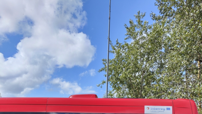 Sacensību uzvarētāju kopbilde. Visi cilvēki tērpušies melnās sporta drēbēs. Divi vīrieši un viena sieviete tur rokās katrs savu izcīnīto zelta kausu. Blakus esošiem sacensību dalībniekiem ap kaklu apliktas iegūtās medaļas. Fonā sarkanas krāsas mikroautobuss