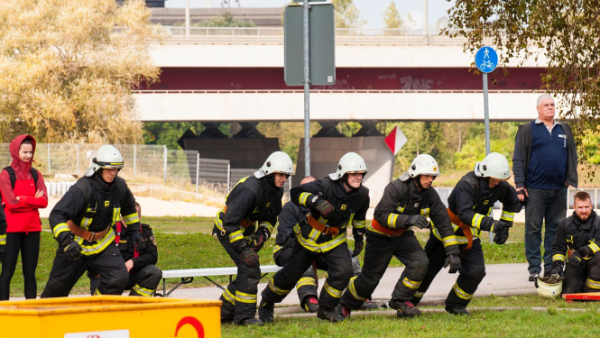 Pieci vīrieši, tērpušies ugunsdzēsēju glābēju aizsargtērpā, gatavojas skriet pa zaļo zālienu. Viņu tuvumā stāv daži skatītāji.