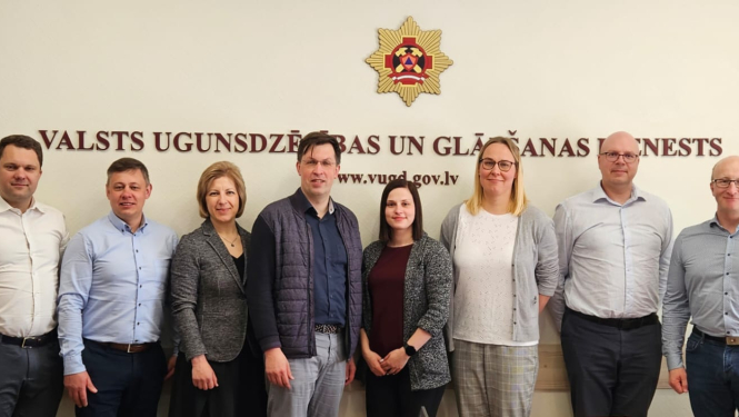 Pieci vīrieši un trīs sievietes vienā rindā stāv un skatās fotogrāfa virzienā. Fonā uz sienas redzama VUGD emblēma un uzraksts bordo krāsā: Valsts ugunsdzēsības un glābšanas dienests