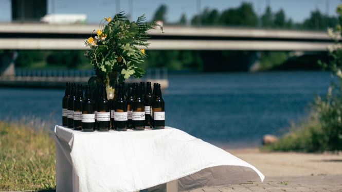 Uz maza galdiņa, kas apklāts ar baltu galdautu, novietotas tumši brūnas atvērtas alus pudeles un jāņuzāļu pušķis. Alus pudelēm uzlīmētas etiķetes ar aicinājumu aizdomāties par to, lai svētkus pavadītu droši