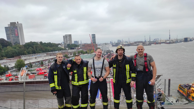 VUGD sportistu - piecu vīriešu - kopbilde. Trīs vīrieši tērpušies ugunsdzēsēju glābēju drēbēs.