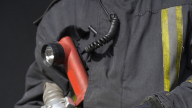 Ugunsdzēsējs glābējs, tērpies aizsargdrēbēs, tur rokās ūdens šļūtenes stobru.