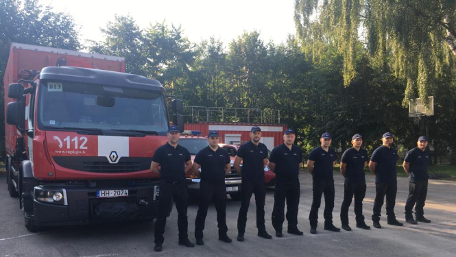 Astoņi vīrieši, tērpušies formastērpos, stāv vienā taisnā līnijā viens otram blakus un skatās fotogrāfa virzienā. Aiz viņiem novietoti VUGD sarkanbaltie transportlīdzekļi