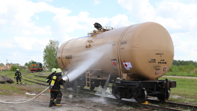 Uz dzelzceļa sliedēm stāv dzeltena cisterna. Tai blakus atrodas divi ugunsdzēsēji glābēji, kas uz cisternas ar šļūtenēm lej ūdeni