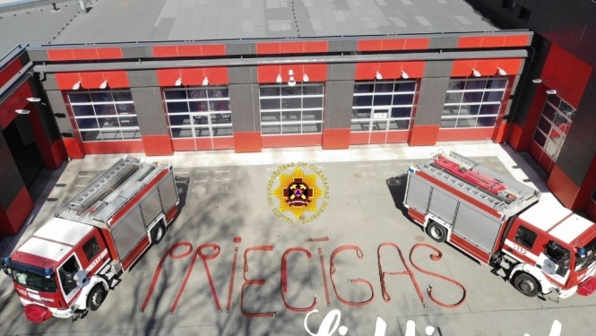 Apsveikums Lieldienās. Tajā redzamas divas ugunsdzēsēju glābēju mašīnas. Mašīnām pa vidu uz zemes ar šļūtenēm izveidots vārds - Priecīgas. Klāt pierakstīts vārds Lieldienas!