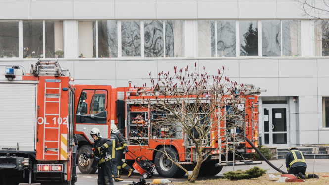 Divas Valsts ugunsdzēsības un glābšanas dienesta mašīnas stāv pie baltas ēkas. Ap mašīnām rosās ugunsdzēsēji glābēji.