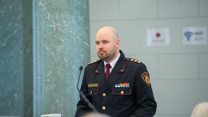 Vīrietis, tērpies dienesta formastērpā, stāv pie galdiņa ar mikrofonu auditorijas priekšā. 