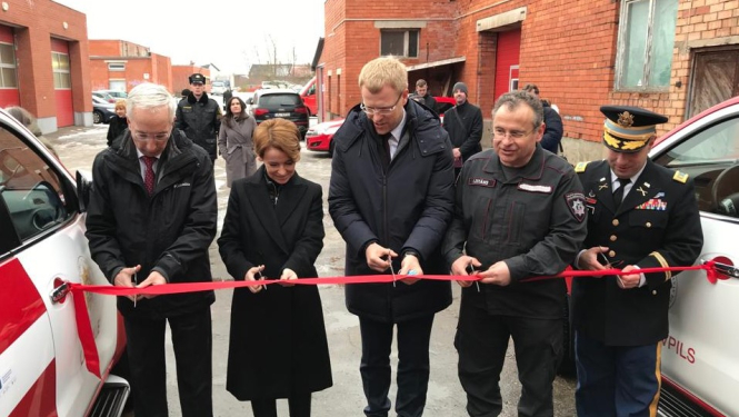 Pieci cilvēki stāv pie sarkanas lentes, kas novilkta no viena ugunsdzēsēju glābēju operatīvā transportlīdzekļa uz otru. Cilvēkiem rokās šķēres. Viņi grasās pārgriezt sarkano lenti.