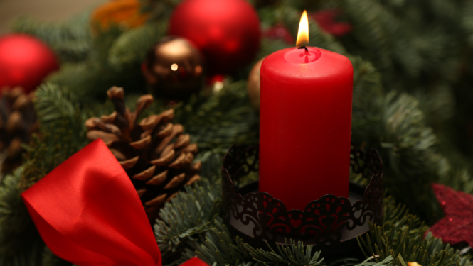 Adventes vainagā aizdegta viena sarkana svece. Netālu no tās atrodas brūns čiekurs, sarkana bantīte, kā arī citi dekoratīvie elementi.