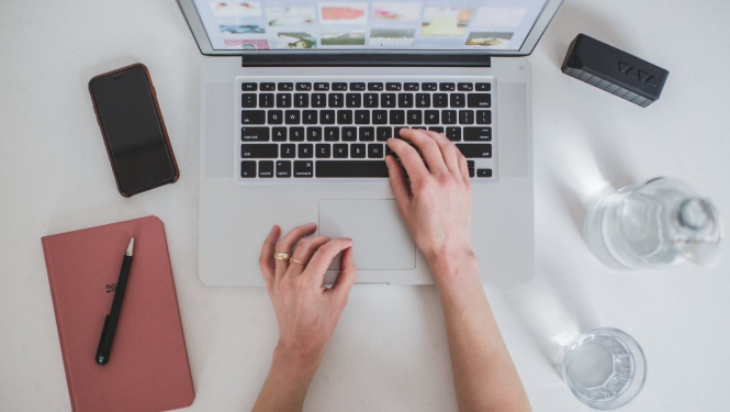 Uz balta galda novietots atvērts portatīvais dators. Tā cilvēks ar savu labo roku pieskaras klaviatūrai, savukārt kreisā roka novietota uz galda. Datora kreisajā malā novietots mobilais tālrunis un rozīgas krāsas blociņš uz kura novietota pildspalva. Datoram labajā malā novietota stikla ūdens pudele un glāze. 