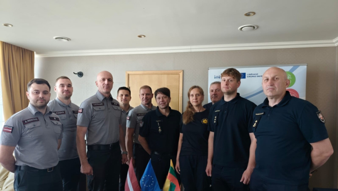 Desmit Latvijas un Lietuvas operatīvo dienestu nodarbinātie stāv ap mazu galdiņu uz kura novietoti trīs karogi - Latvijas, Igaunijas un Lietuvas