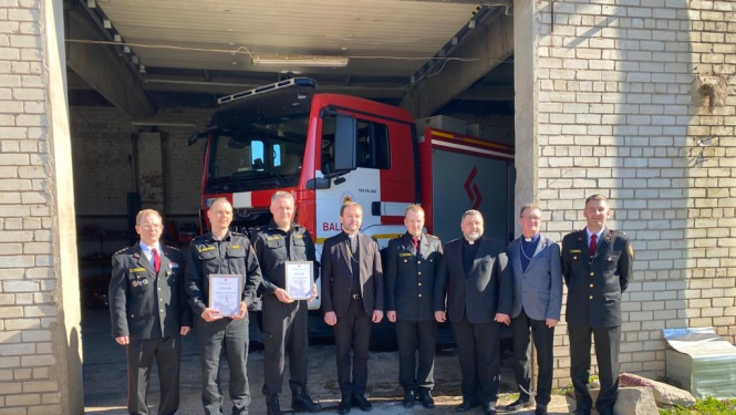 Pie ēkas ar vaļējām durvīm stāv astoņi vīrieši - ugunsdzēsēji glābēji un Baldones baznīcas pārstāvji. Fonā redzama ugunsdzēsēju glābēju automašīna