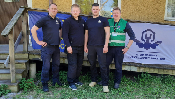 Četri vīrieši stāv zālienā. Fonā redzama dzeltena ēkas siena, kāpnes