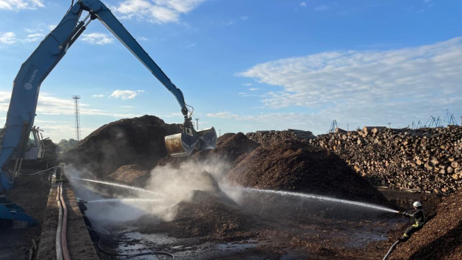 Liels eskavators virs šķeldas kaudzes tur savus kausus. No trīs pusēm ugunsdzēsēji glābēji dzēš gruzdošo šķeldu. Fonā zilas debesis ar baltiem mākonīšiem