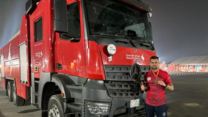 Vīrietis, tērpies tumši sarkanā kreklā un melnās biksēs, stāv pie lielas sarkanas ugunsdzēsēju glābēju automašīnas un tur rokās ap kaklu uzlikto iegūto bronzas medaļu