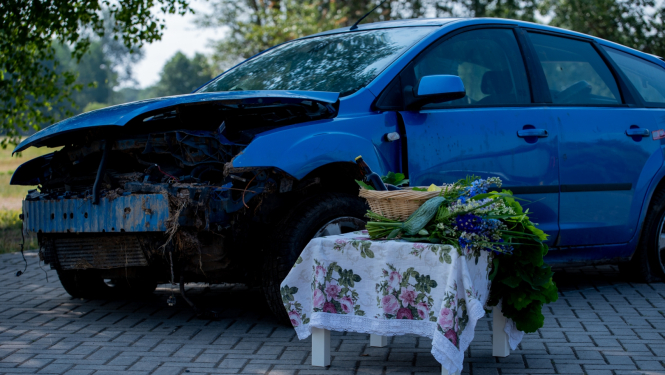 Stāv zila, sasista automašīna. Tai priekšā uz balta galdiņa, kas pārklāts ar raibu galdautu, uzliktas jāņuzāles un grozs ar alkoholu, kā arī svētku gardumiem