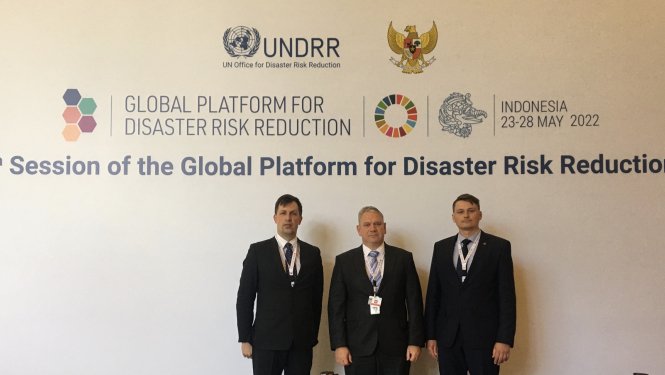 Trīs Valsts ugunsdzēsības un glābšanas pārstāvji stāv pie sienas, uz kuras redzami dažādi pasākuma logo