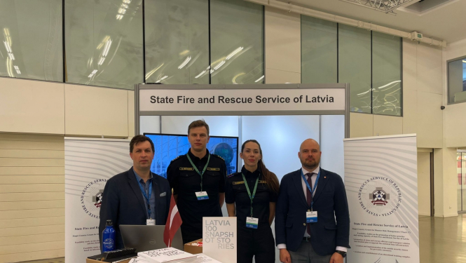 Četri VUGD darbinieki - trīs vīrieši un viena sieviete stāv pie galdiņa uz kura izvietoti dažādi uzskates materiāli. Fonā redzami divi baneri.