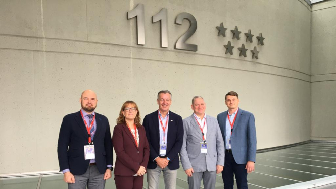 Piecu cilvēku - vienas sievietes un četru vīriešu - kopbilde. Visi stāv un skatās fotogrāfa virzienā. Fonā redzama siena uz kuras ir redzams tālruņa numurs 112 un septiņas zvaigznes.