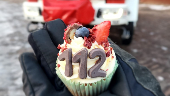 Redzamas cilvēka plaukstas, kas ietērptas melnos cimdos. Tajās tiek turēts mazs konditorejas izstrādājums ar cipariem 112. Fonā novietota ugunsdzēsēju glābēju autocisterna