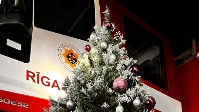 Ziemassvētku eglīte ar rotājumiem. Tai pie kājām novietota ugunsdzēsēju glābēju jaka, bet aiz eglītes atrodas ugunsdzēsēju glābēju autocisterna