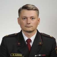 Vīrietis, tērpies tumšas krāsas formastērpā, skatās fotogrāfa virzienā.