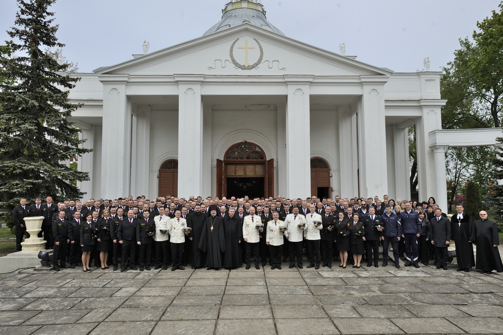 Pie baltas baznīcas stāv formastērpos tērpušies VUGD nodarbinātie un svinību viesi. Visi skatās fotogrāfa virzienā.
