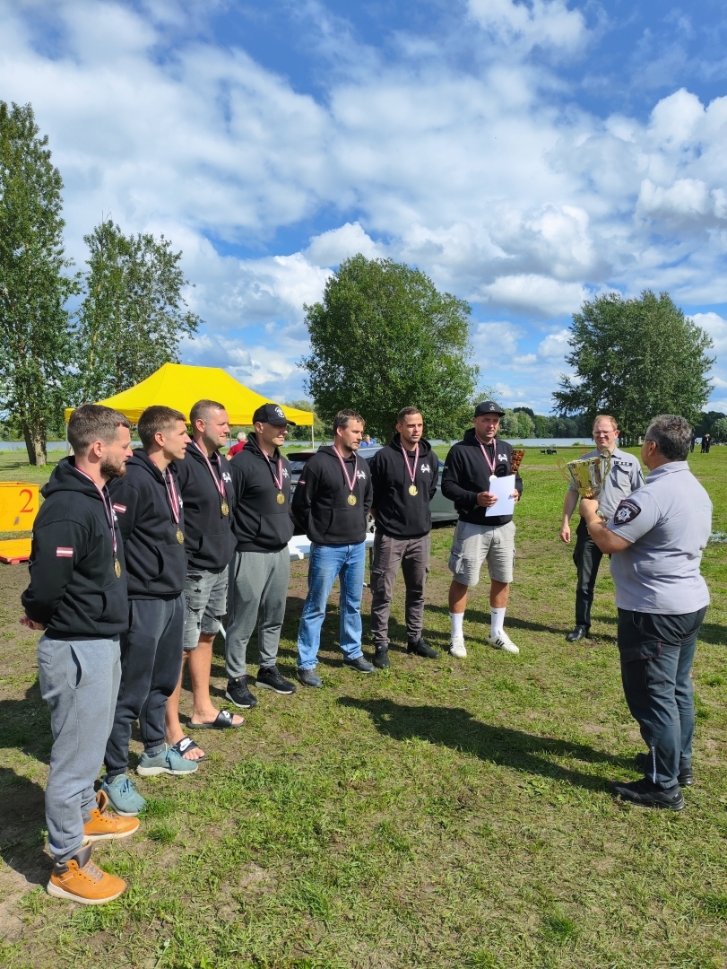 Septiņi vīrieši, tērpušies sporta tērpos, stāv ierindā un skatās uz viņiem pretim stāvošo VUGD nodarbināto. Visiem septiņiem vīriešiem kaklā ir uzkārtas iegūtās sacensību medaļas. Viens no vīriešiem rokās tur arī iegūto diplomu