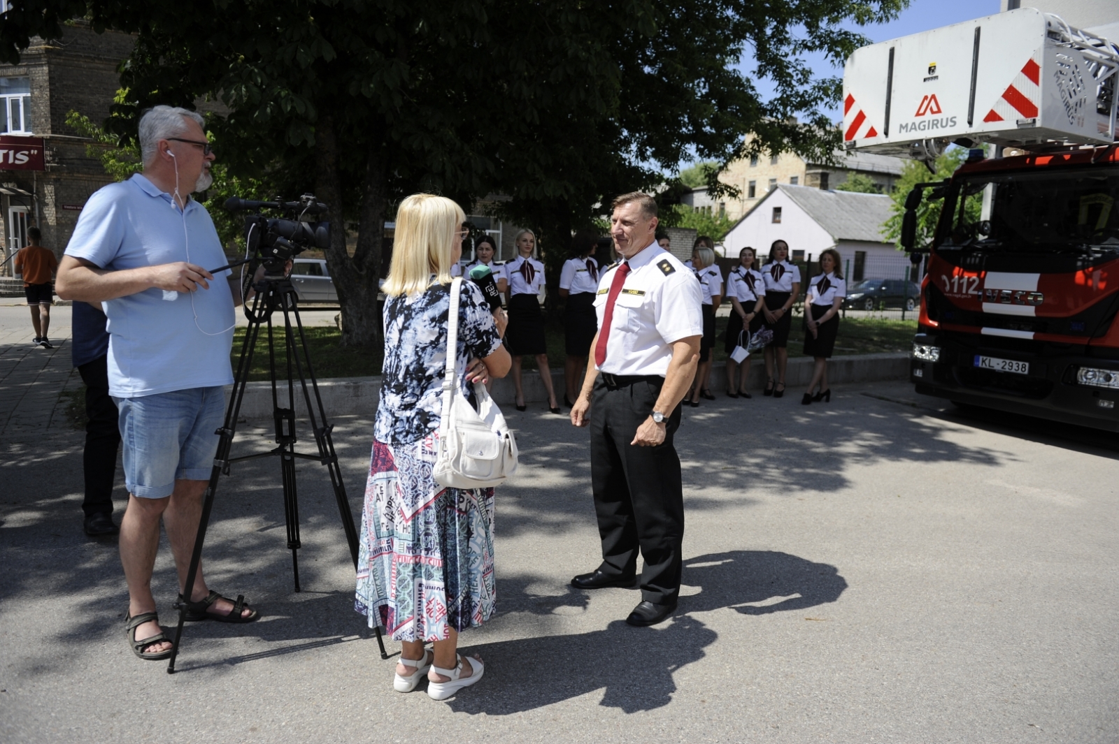 VUGD nodarbinātais sarunājas ar žurnālisti, kas tērpusies raibā kleitā. Viņai blakus stāv operators ar video kameru, kas novietota uz melna statīva.