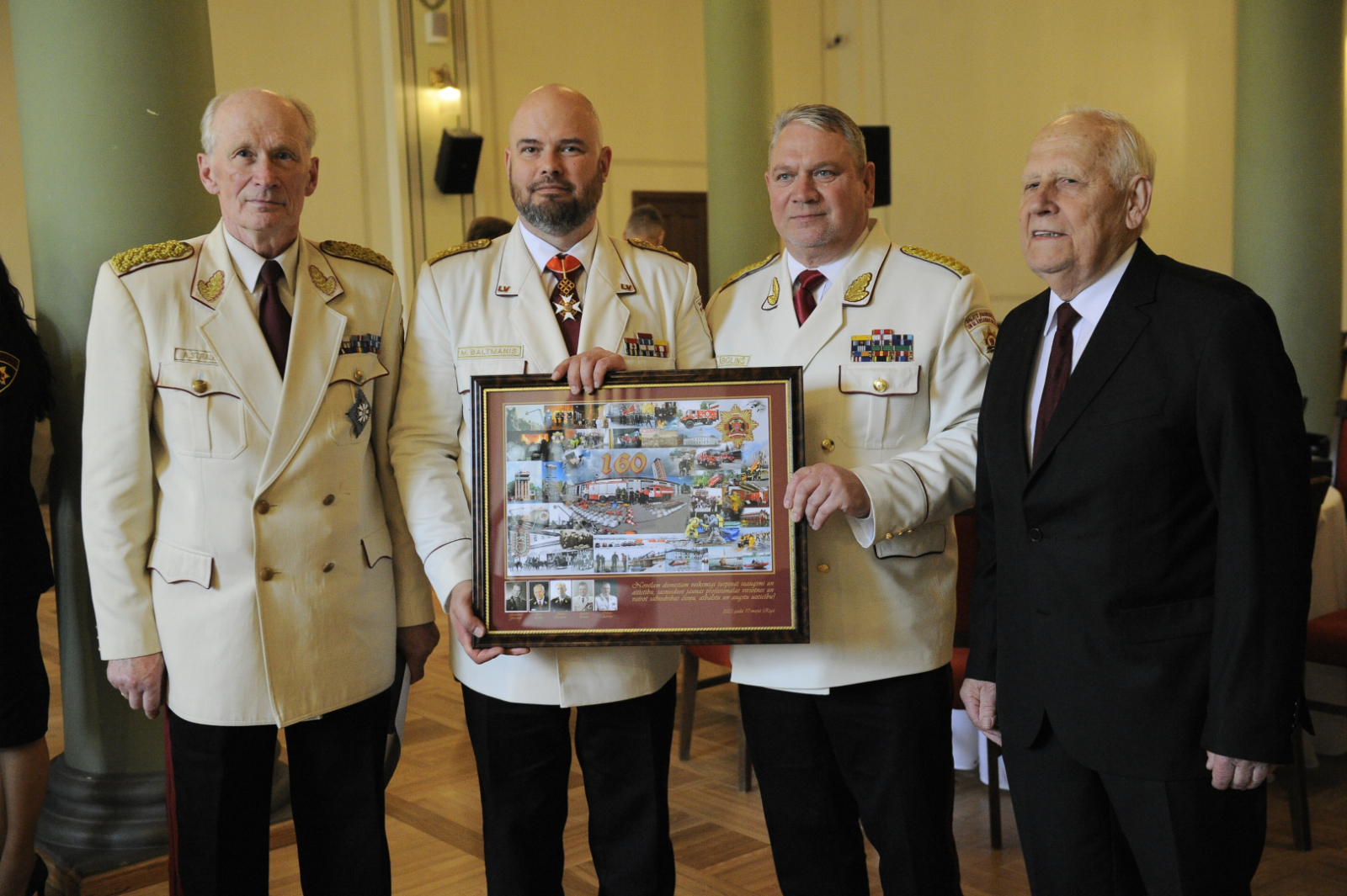 Četri svinīgos tērpos tērpti vīrieši stāv un skatās fotogrāfa virzienā. Divi no viņiem tur vienu ierāmētu plakātu. 