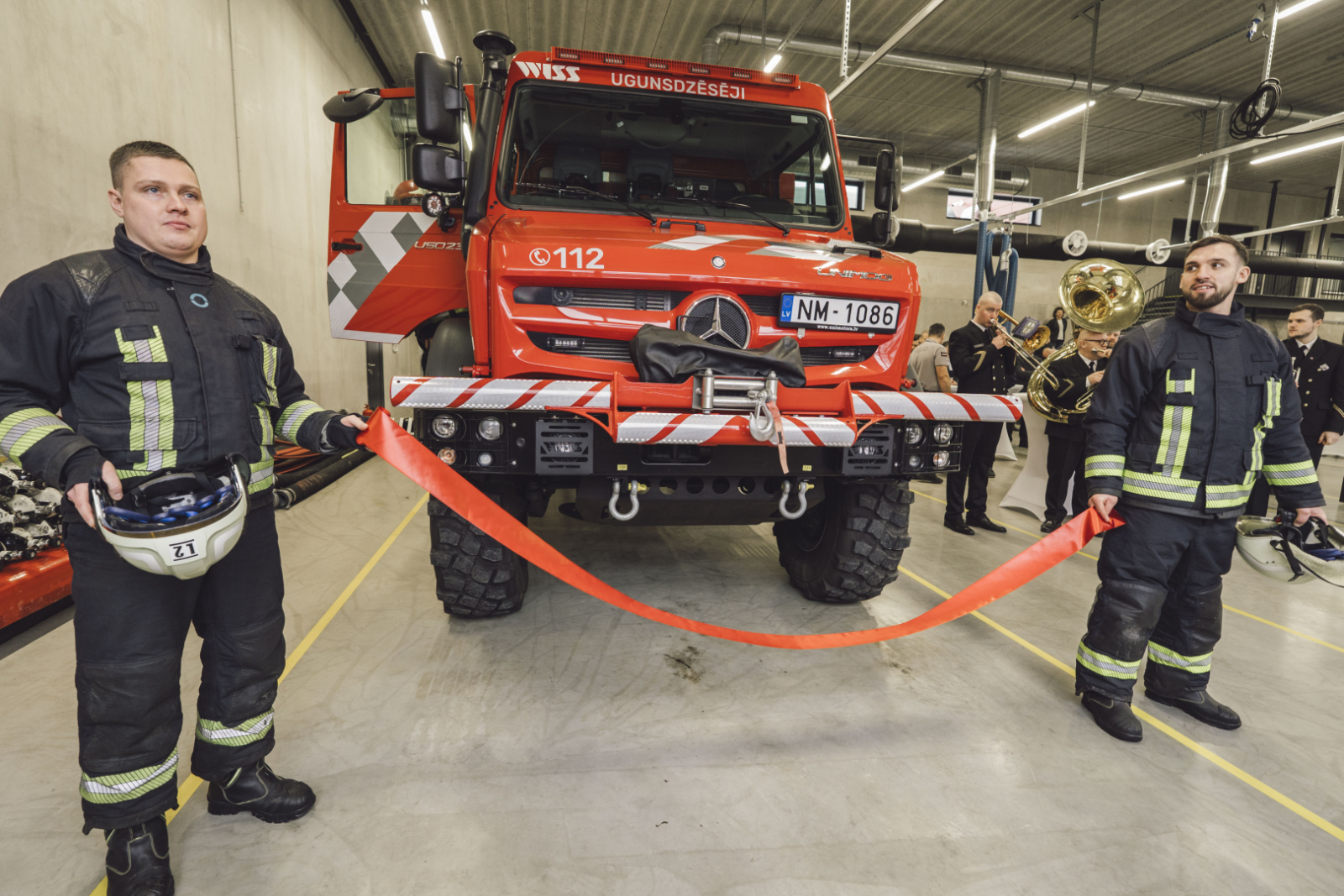Fonā VUGD specializētais transportlīdzeklis. Priekšplānā divi ugunsdzēsēji glābēji tur sarkanu lenti.