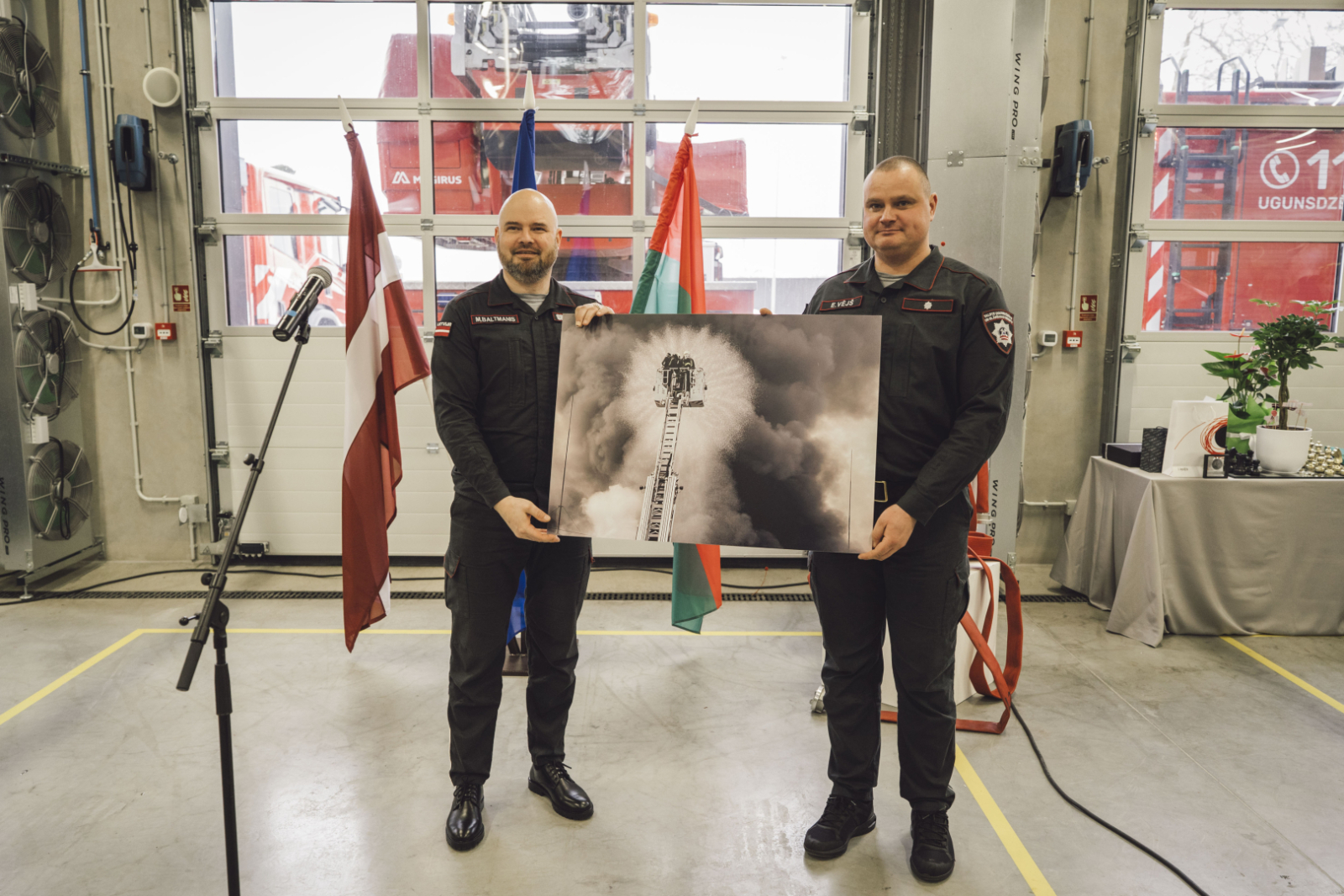 Valsts ugunsdzēsības un glābšanas dienesta priekšnieks ģenerālis Mārtiņš Baltmanis un Liepājas 2. daļas komandieris Edgars Vējš tur lielizmēra fotogrāfiju, kurā attēloti ugunsdzēsēji glābēji.