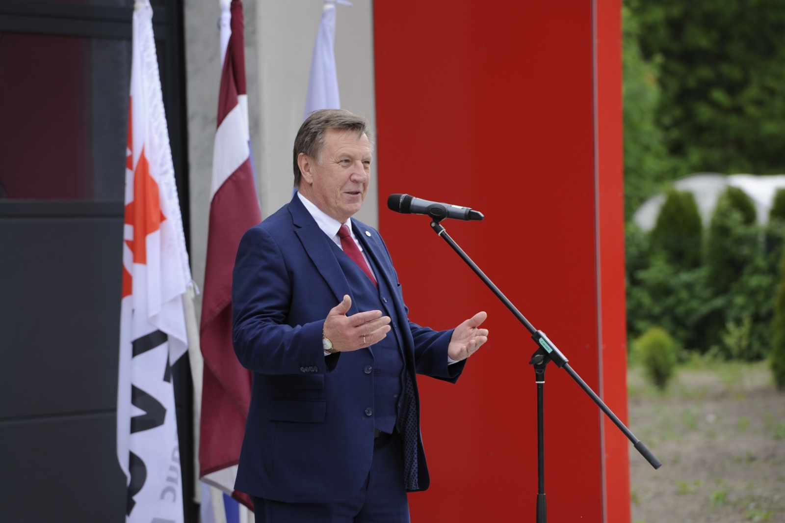 Iekšlietu ministris Māris Kučinskis, tērpies zilā uzvalkā, stāv pie mikrofona un saka uzrunu. Fonā trīs karogi. Viens no tiem ir Latvijas valsts karogs.