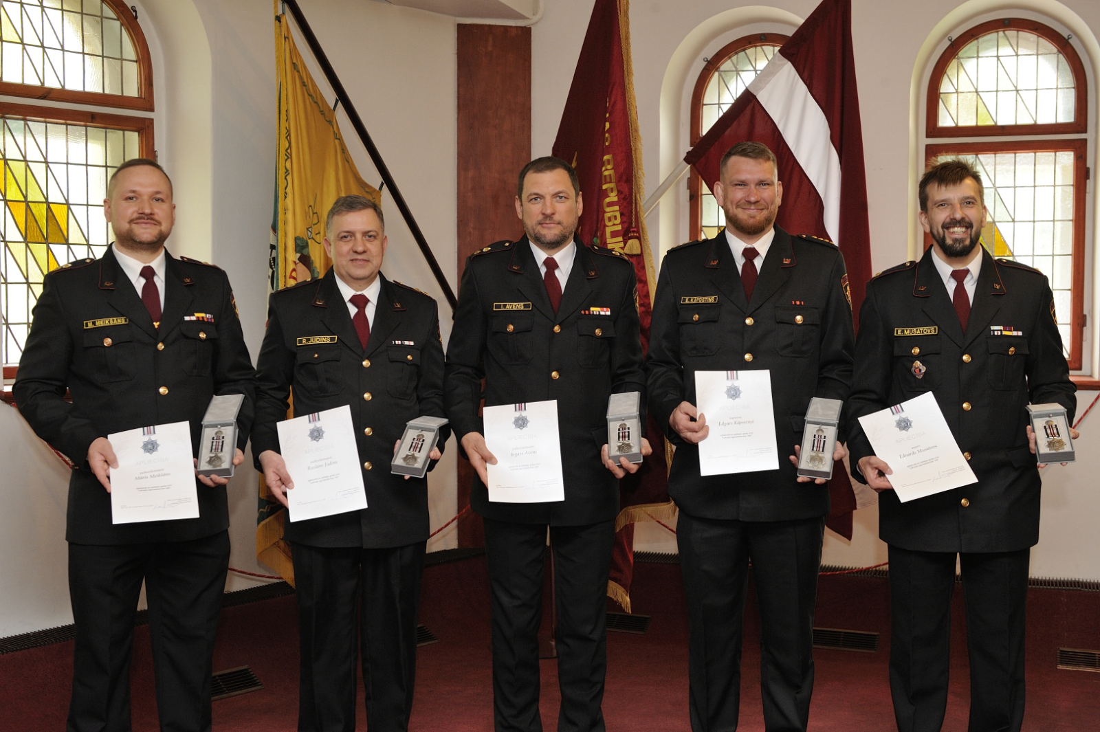 Pieci formastērpos tērpušies vīrieši stāv, rokās turot iegūtos apbalvojuma rakstus un medaļas. Aiz viņiem izvietoti trīs karogi, tostarp Latvijas ugunsdzēsības muzeja un Latvijas.