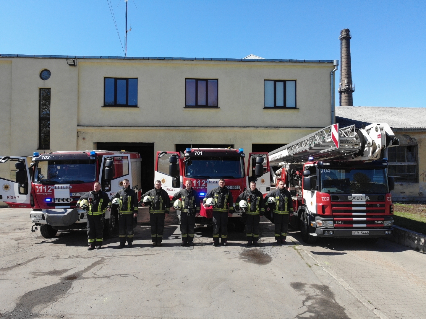 Septiņi ugunsdzēsēji glābēji stāv, turot rokās savas ķiveres. Viņiem fonā redzamas trīs dažādas ugunsdzēsēju glābēju autotehnikas