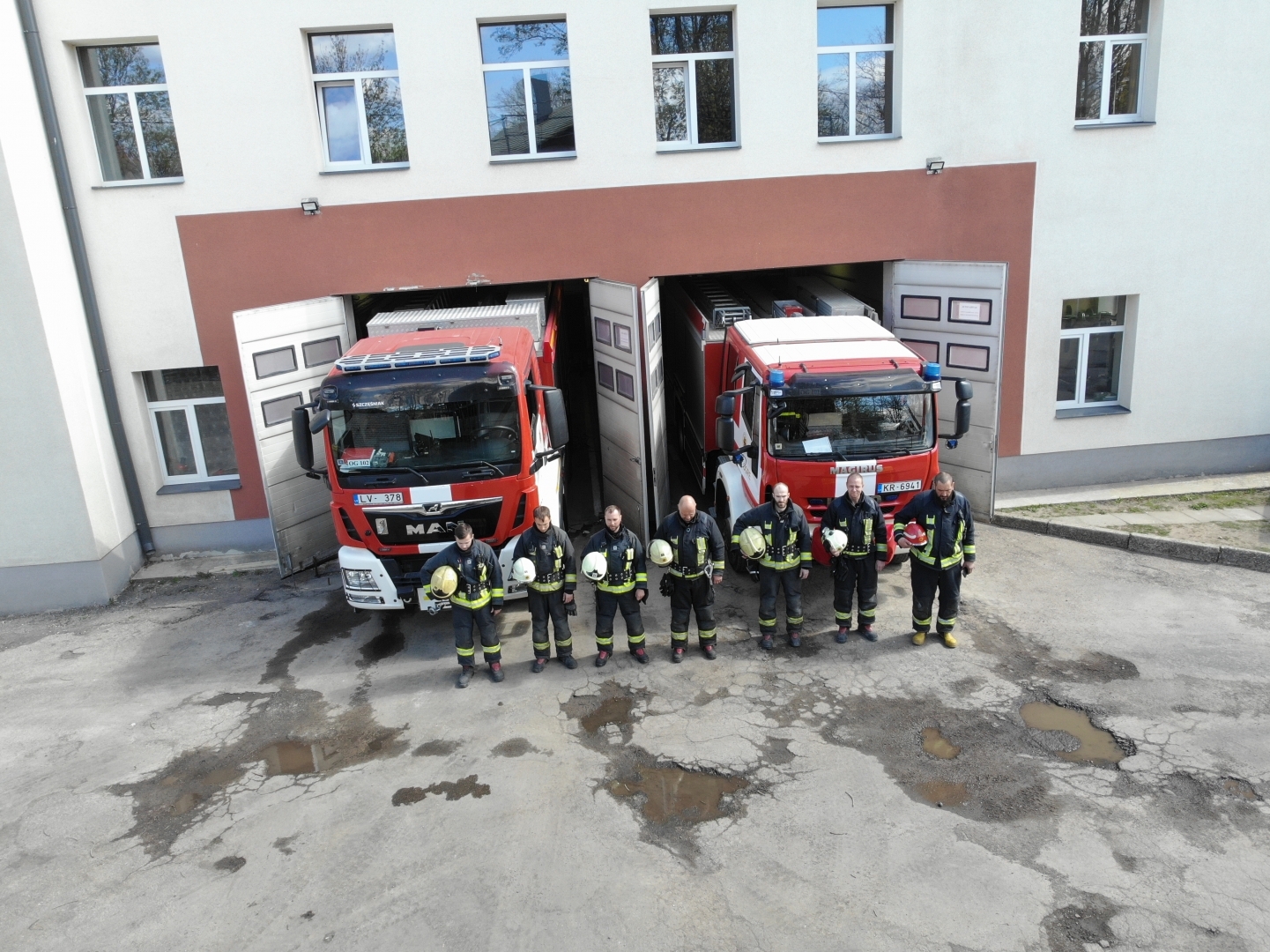 Septiņi ugunsdzēsēji glābēji stāv vienā rindā. Rokās tur savas ķiveres. Aiz viņiem redzamas divas ugunsdzēsēju glābēju automašīnas