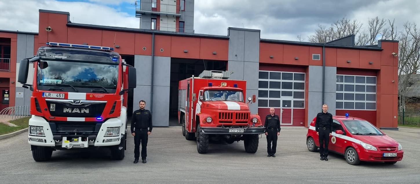 Trīs ugunsdzēsēji glābēji un trīs ugunsdzēsēju glābēju automašīnas. Katrs ugunsdzēsējs glābējs stāv pie savas ugunsdzēsības glābšanas mašīnas