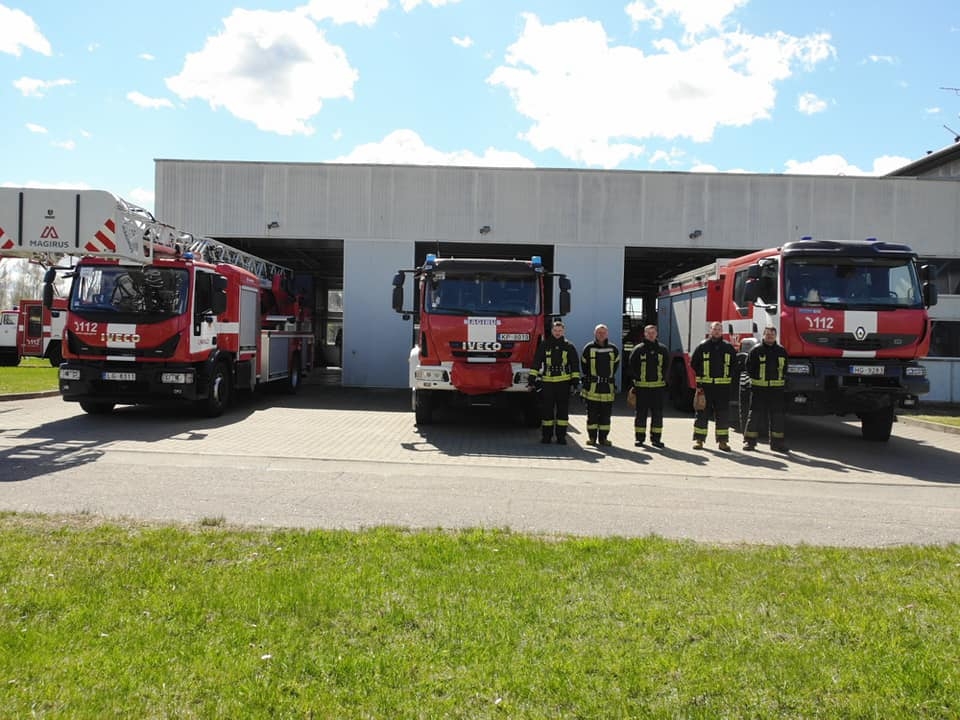Pieci ugunsdzēsēji glābēji stāv, ieturot klusuma brīdi. Viņiem fonā redzamas trīs ugunsdzēsēju glābēju mašīnas