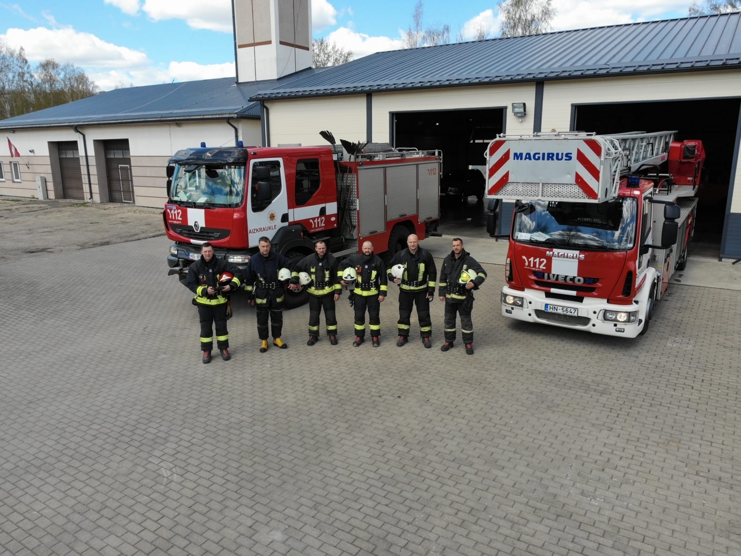 Seši ugunsdzēsēji glābēji stāv vienā rindā un ietur klusuma brīdi. Viņiem fonā redzamas divas ugunsdzēsēju glābēju mašīnas