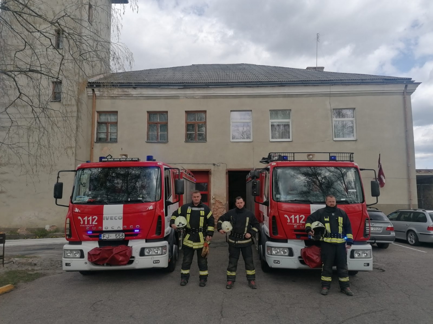 Trīs ugunsdzēsēji glābēji ietur Klusuma brīdi. Viņiem aiz muguras ir redzamas divas ugunsdzēsēju glābēju mašīnas