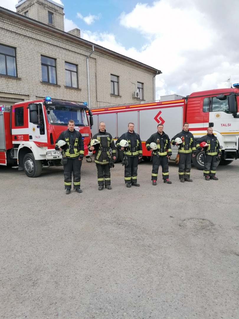 Seši ugunsdzēsēji glābēji stāv vienā rindā, tur rokās ķiveres un ietur Klusuma brīdi. Viņiem aiz muguras redzamas divas ugunsdzēsēju glābēju mašīnas