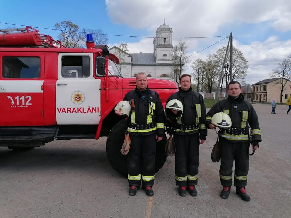 Trīs ugunsdzēsēji glābēji stāv pie vienas ugunsdzēsēju glābēju mašīnas un ietur Klusuma brīdi