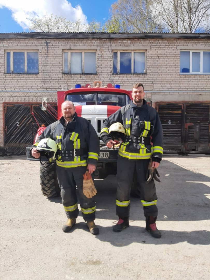 Ģērbušies savos aizsargtērpos un turot rokās ķiveri stāv divi ugunsdzēsēji glābēji. Viņiem fonā redzama ugunsdzēsības un glābšanas dienesta automašīna