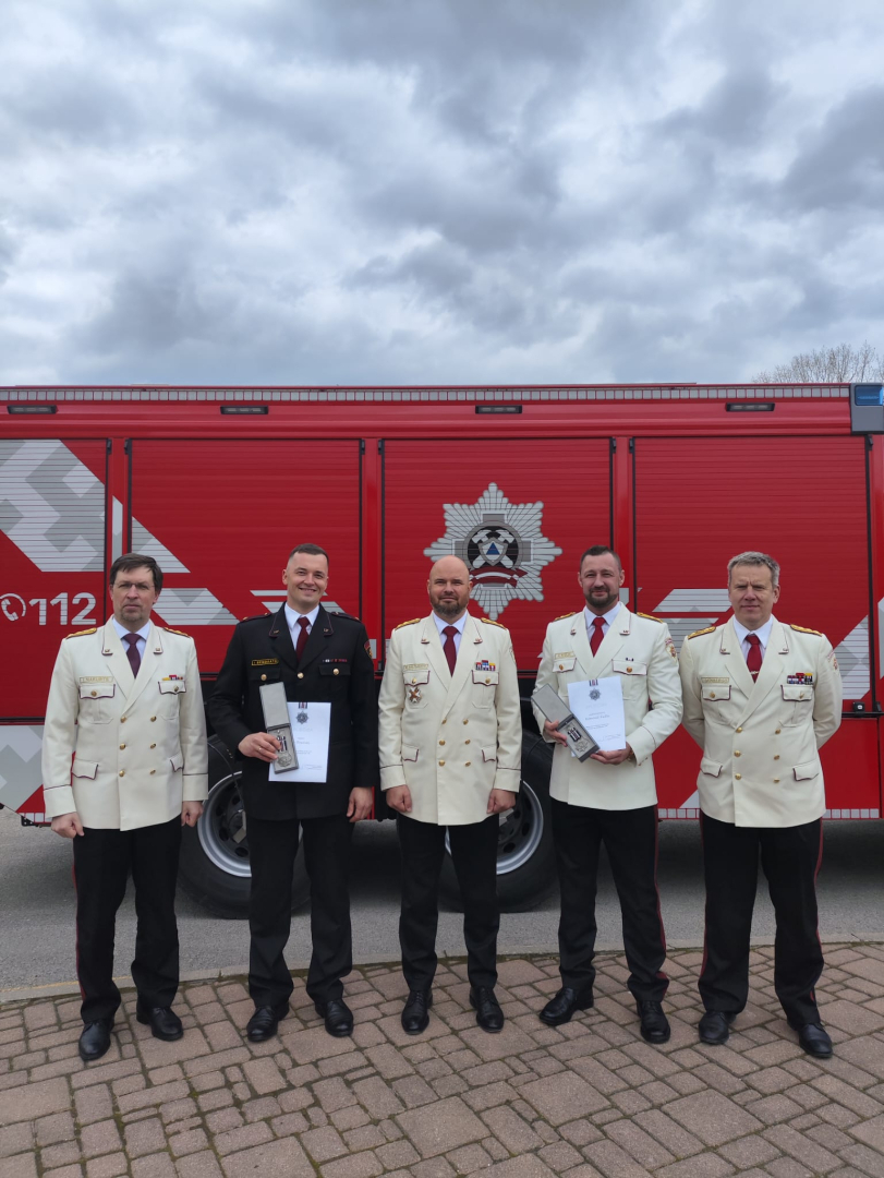Pieci vīrieši, kas tērpušies savos svinīgajos formastērpos, skatās fotogrāfa virzienā. Divi no viņiem rokās tur iegūtos apbalvojumus. Viņiem fonā stāv ugunsdzēsēju glābēju sarkanā autocisterna.