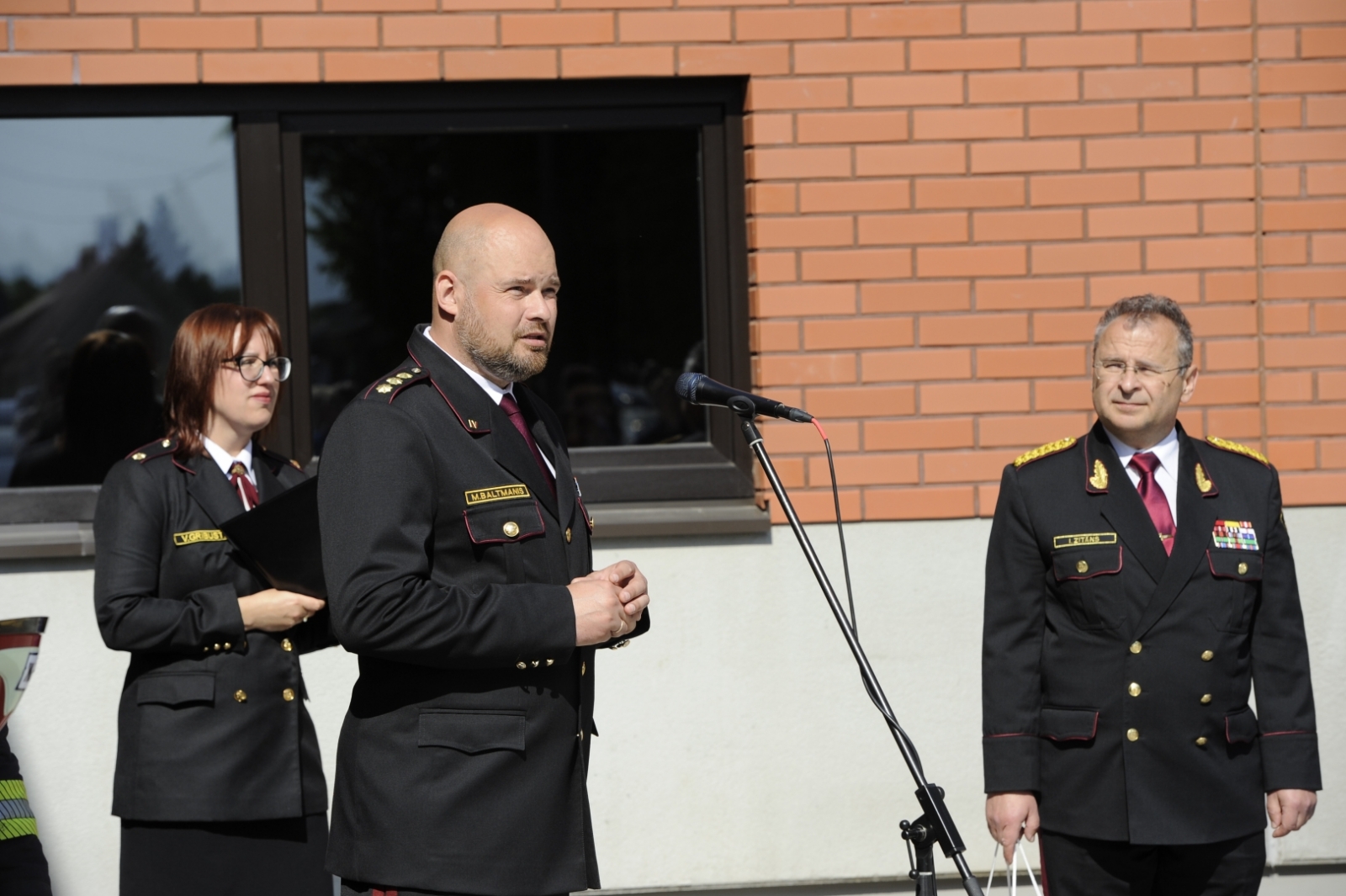 VUGD priekšnieks Mārtiņš Baltmanis stāv pie mikrofona un saka uzrunu. Fonā redzama pelēki sarkanīgā depo daļa un divi VUGD nodarbinātie.