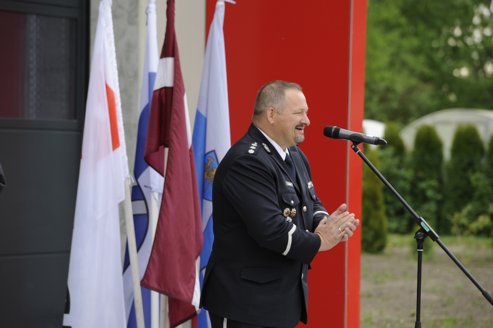 Policijas priekšnieks stāv pie mikrofona un saka uzrunu. Uzrunas brīdī roku plaukstas ir salicis kopā. Fonā trīs karogi un VUGD depo.