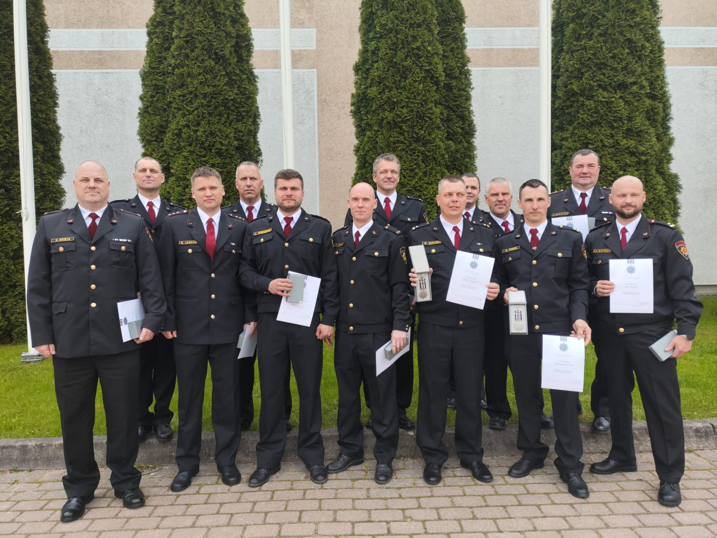 Vairāki formastērpos tērpti vīrieši stāv un skatās fotogrāfa virzienā, rokās turot iegūtos apbalvojumus. Viņiem fonā redzamas zaļas tūjas, aiz kurām bēšīgas krāsas ēkas siena.