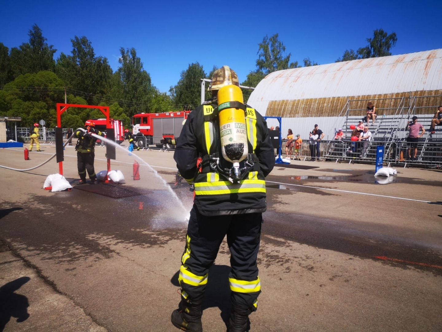 Ugunsdzēsējs glābējs, tērpies pilnā ekipējumā un vēl ar gāzes masku un balonu, skatās, kā otrs ugunsdzēsējs glābējs darbojas ar ūdens šļūteni