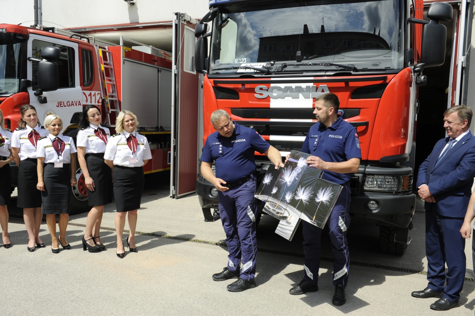 Divi valsts policijas nodarbinātie stāv pie sarkanās autocisternas un tur rokās gleznu - baltas pieneņpūkas uz pelēkas fona. Kreisajā pusē stāv dispečeres, bet labajā iekšlietu ministrs.